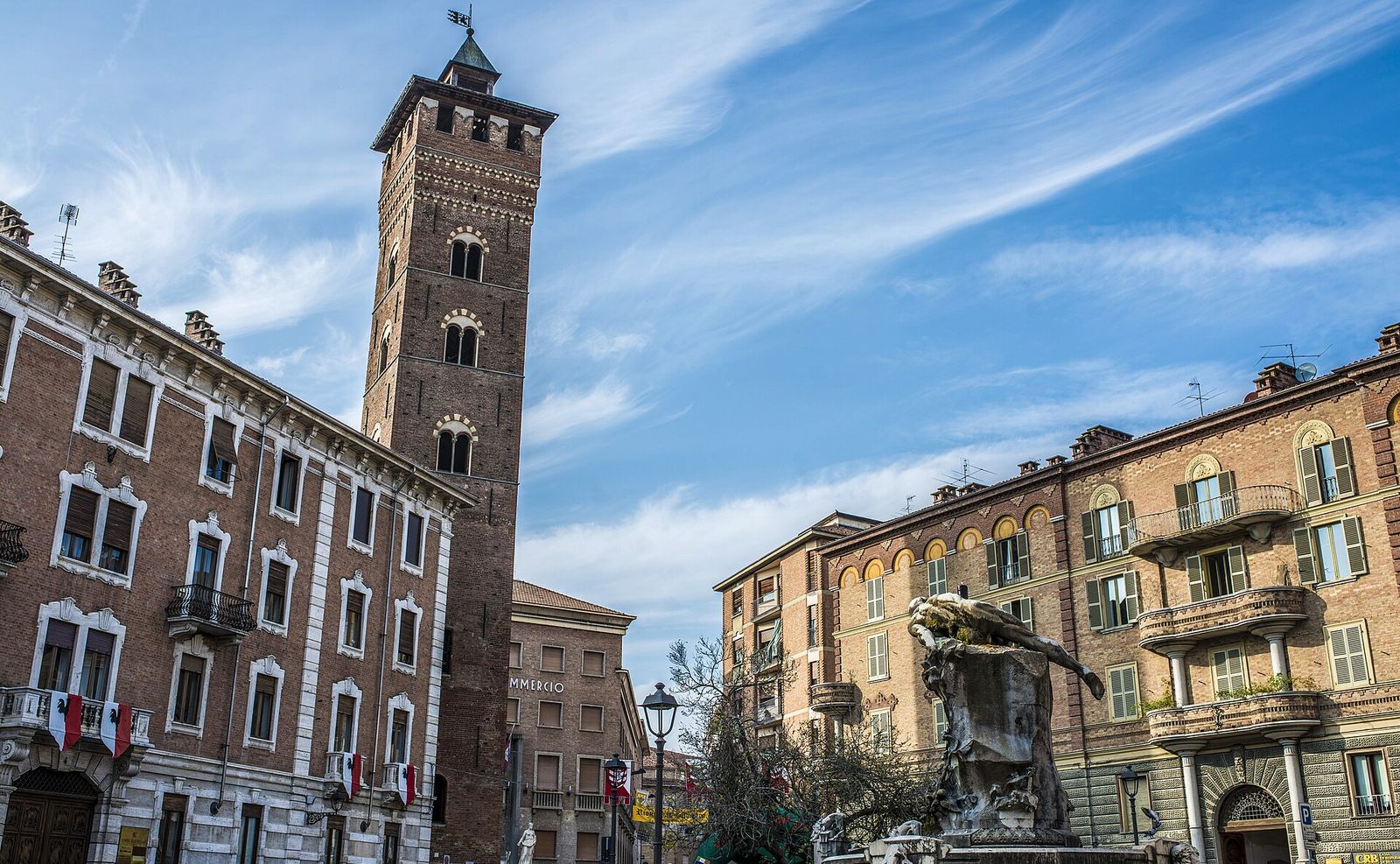 La Torre Troyana in piazza Medici ad Asti, con un gruppo scultoreo in primo piano e bandiere bianche e rosse ai balconi dei palazzi